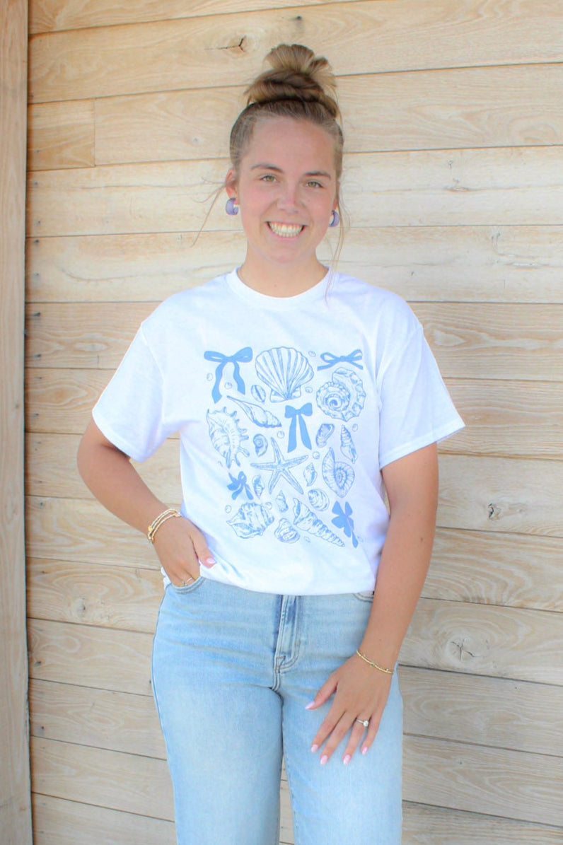 Person wearing a white t-shirt with graphics and light blue jeans standing in front of a wooden building.